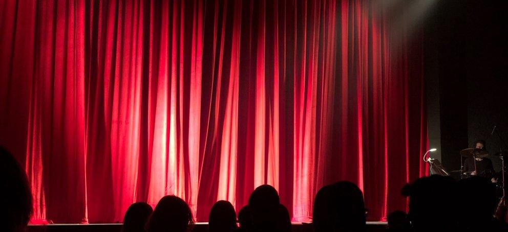 Red curtain lit up by stage lights with shadows of audience in the foreground.